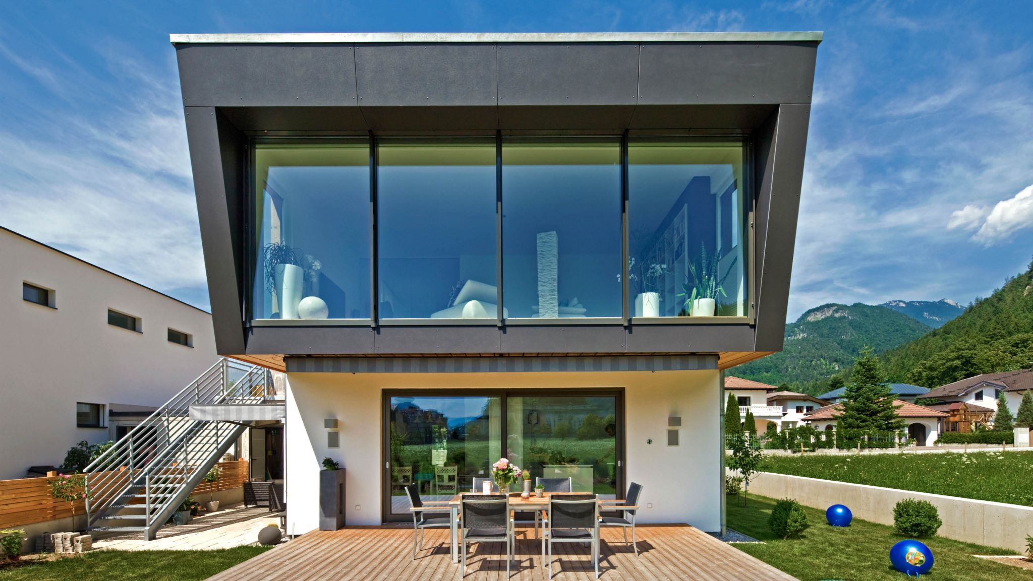 Moderne Architektur: Einblick in ein futuristisches Haus mit Glasfassade, Holzterrasse und Bergblick im Sommer.