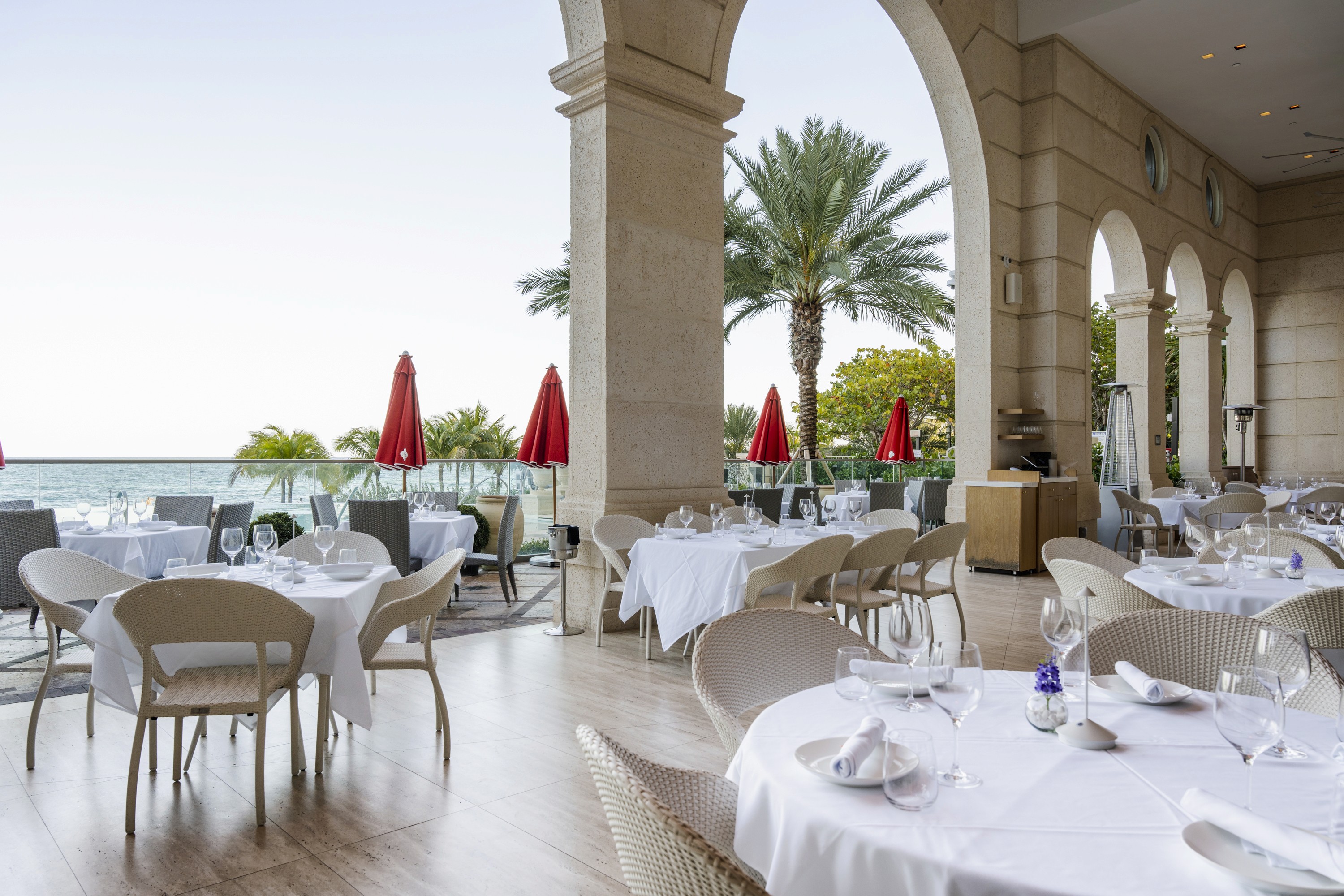 Elegant outdoor restaurant with white tables, palm trees, and red umbrellas, overlooking the ocean.