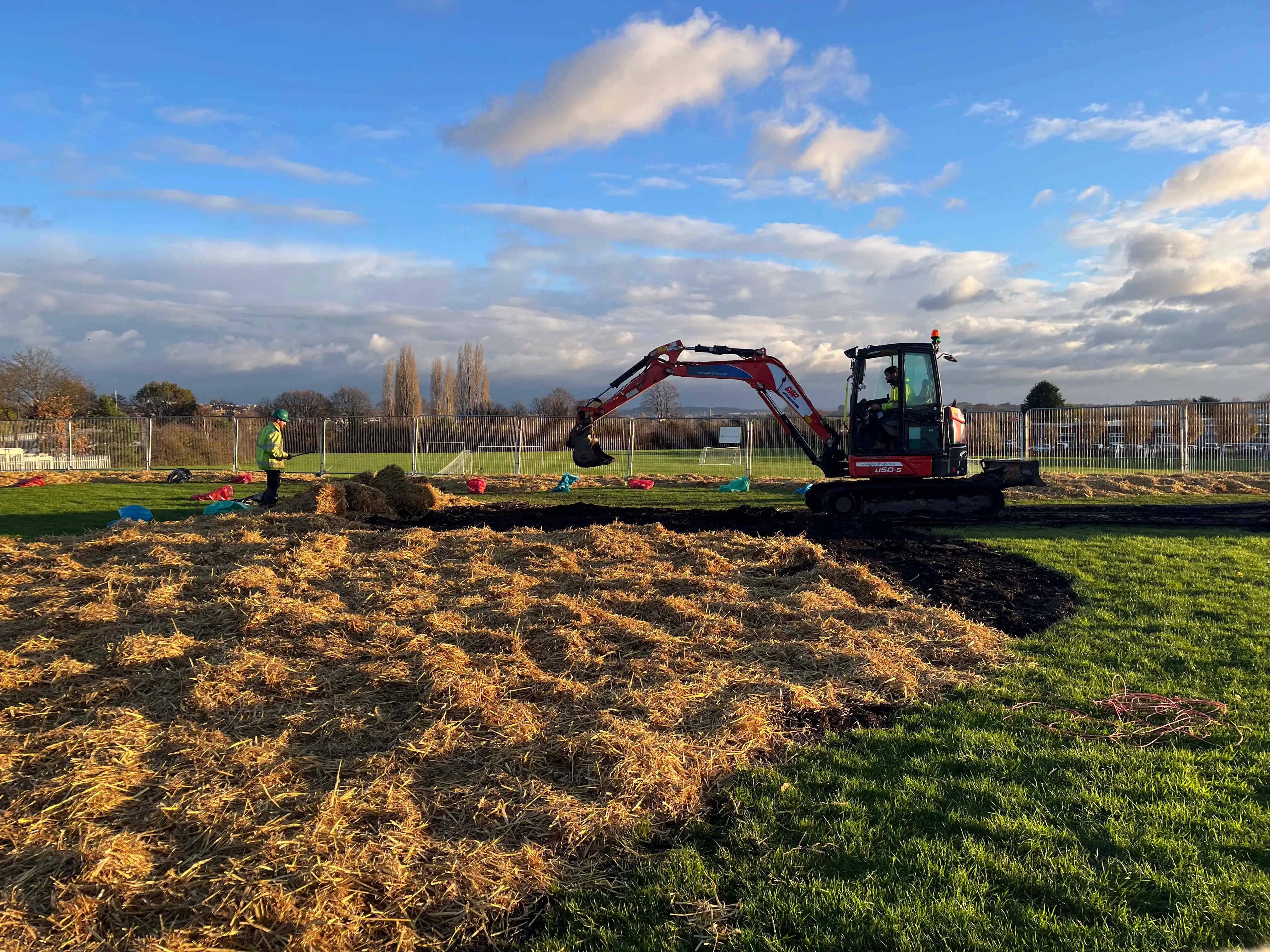 A tractor plows a field under a blue sky with clouds, surrounded by green grass and distant hills.