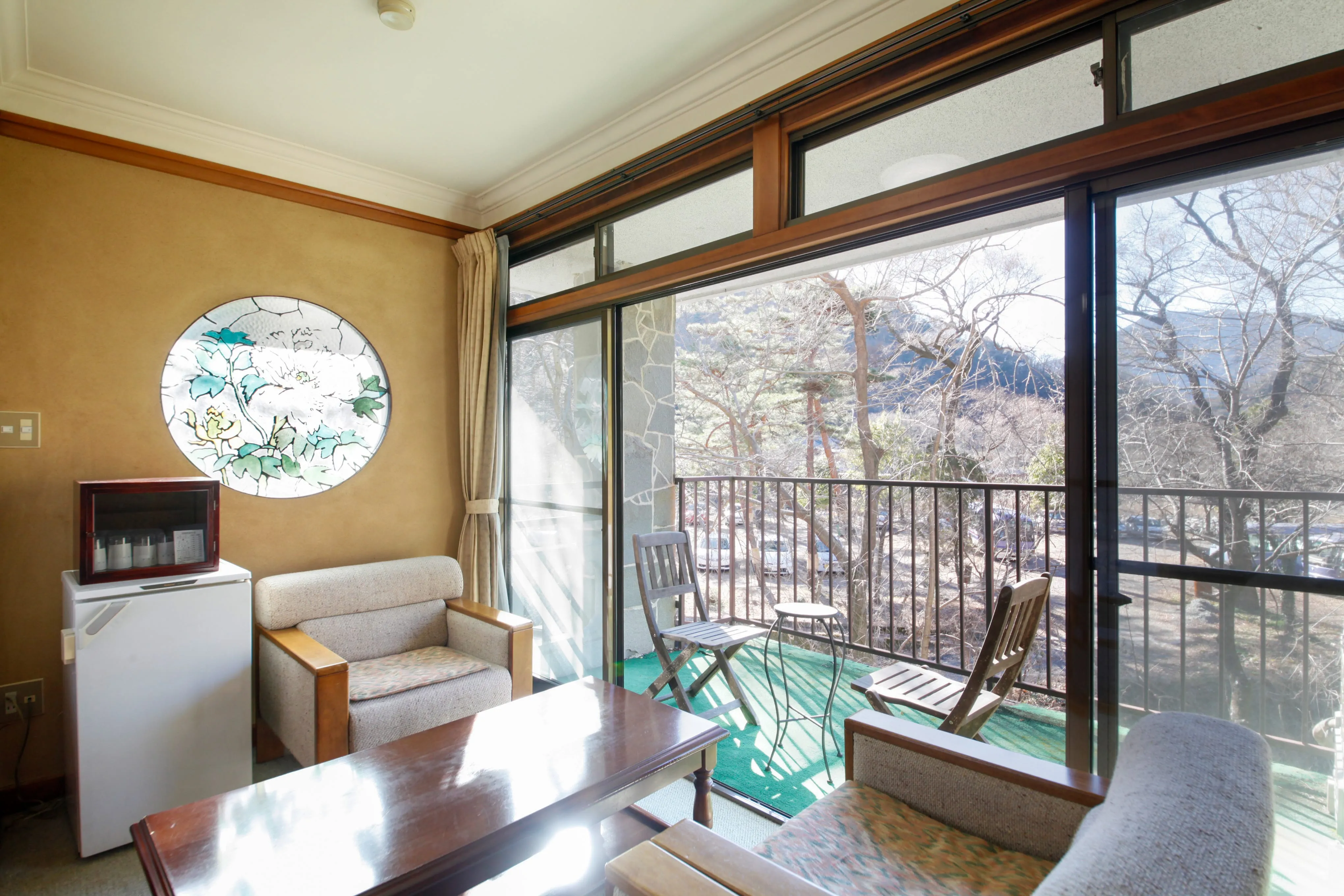 A wide-angle view of a spacious, traditional Japanese interior with tatami mat floors. To the right is a bright living area with a low table, floor cushions, and shoji screen windows. To the left, a more rustic space features a sunken irori hearth with a decorative pot hook hanging from the ceiling.
