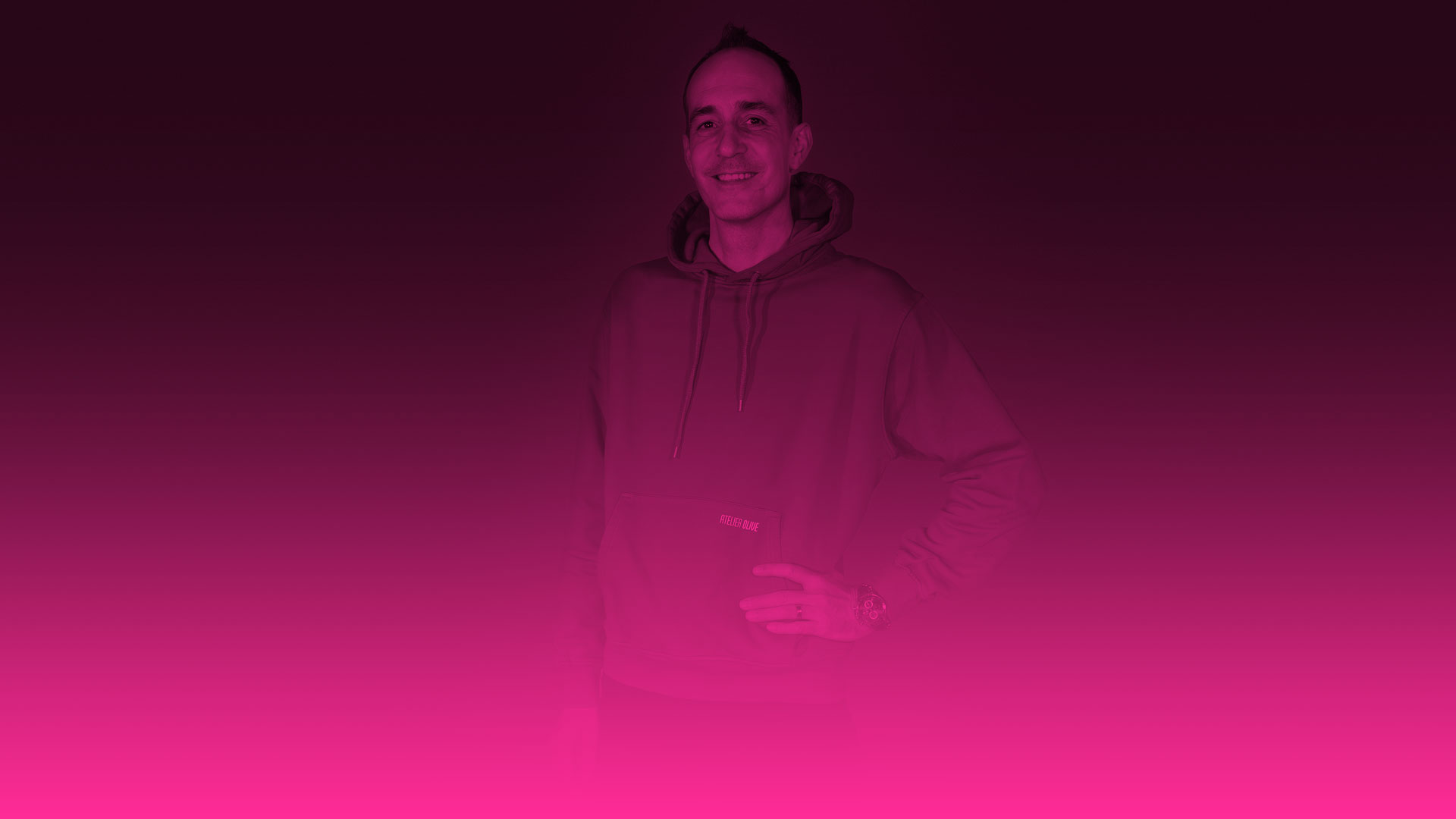 Das Foto zeigt das Porträt eines lächelnden Mannes, der direkt in die Kamera blickt. Er trägt einen sportlichen Kapuzenpullover (Hoodie) und hat eine Hand entspannt in die Hüfte gestützt. Am linken Handgelenk ist eine dunkle Armbanduhr zu erkennen. Das gesamte Bild ist stark verfremdet und mit einem intensiven Farbverlauf belegt: Die obere Hälfte ist in einem dunklen Violett-Ton gehalten, der nach unten hin fließend in ein leuchtendes Magenta (Pink) übergeht. Der untere Bildrand verschmilzt komplett mit einer durchgehenden, leuchtend pinken Farbfläche. Der Hintergrund hinter dem Mann ist dunkel und ohne erkennbare Details.