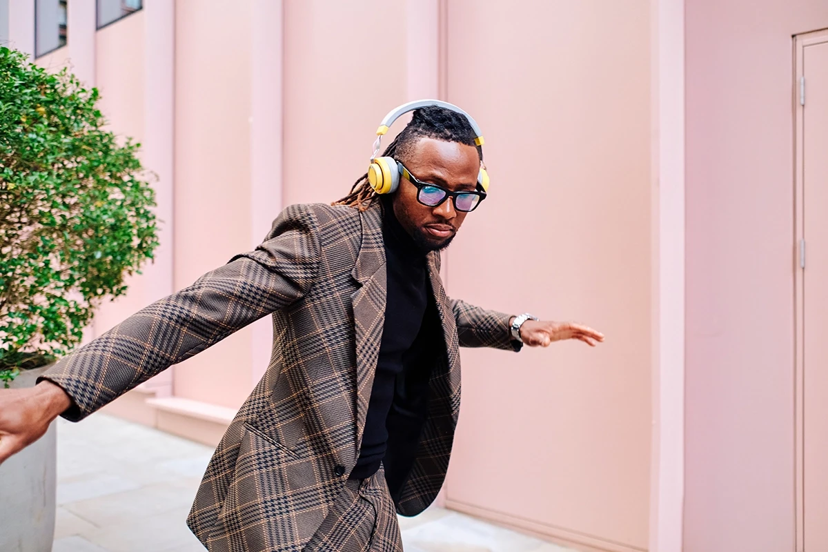 Stylish man dancing outdoors wearing yellow over-ear headphones, plaid blazer, mirrored sunglasses and silver watch against pastel pink wall, urban street style