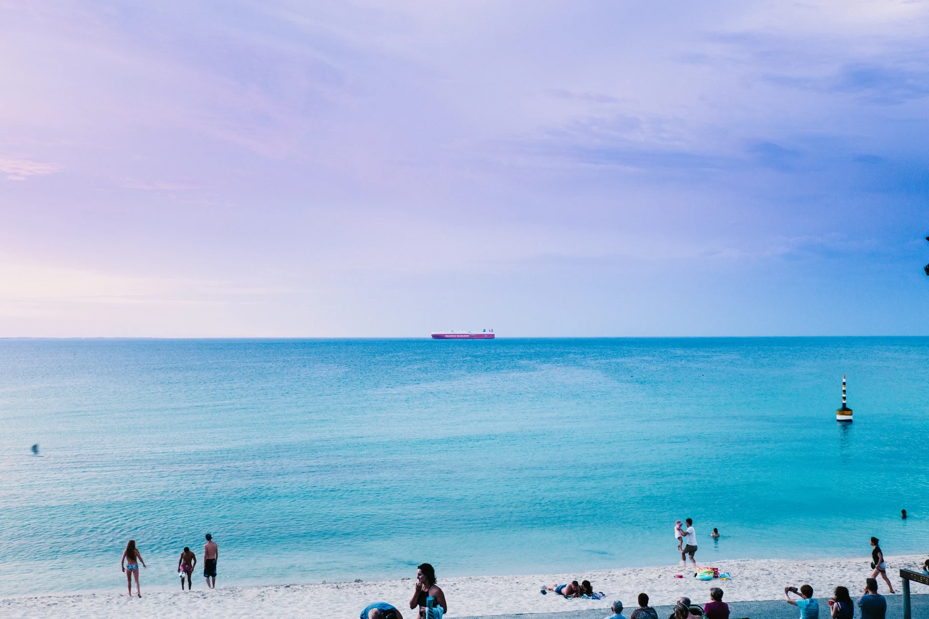 A cargo ship on the horizon beyond calm turquoise water, with beachgoers in the foreground