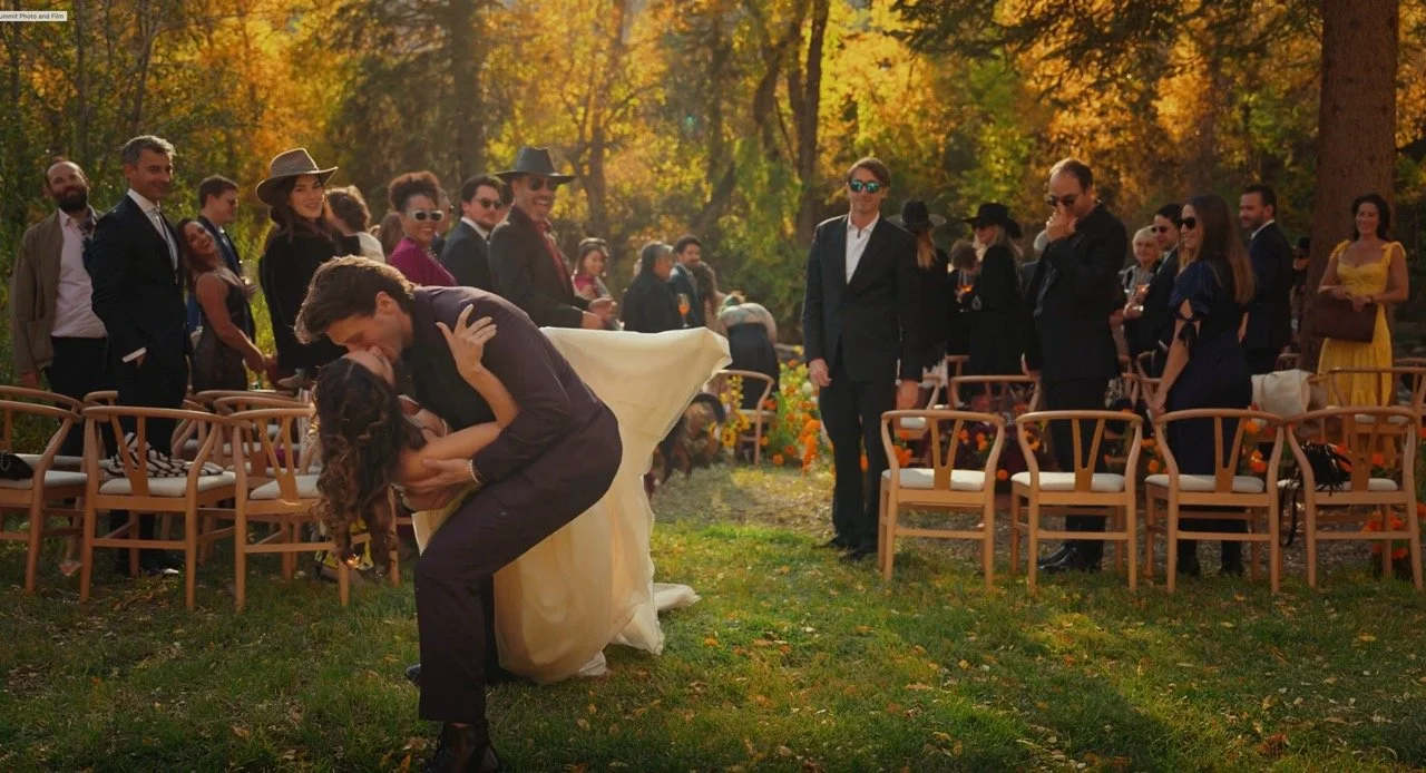 Newlyweds share a dip kiss in the ceremony aisle surrounded by guests and autumn trees.