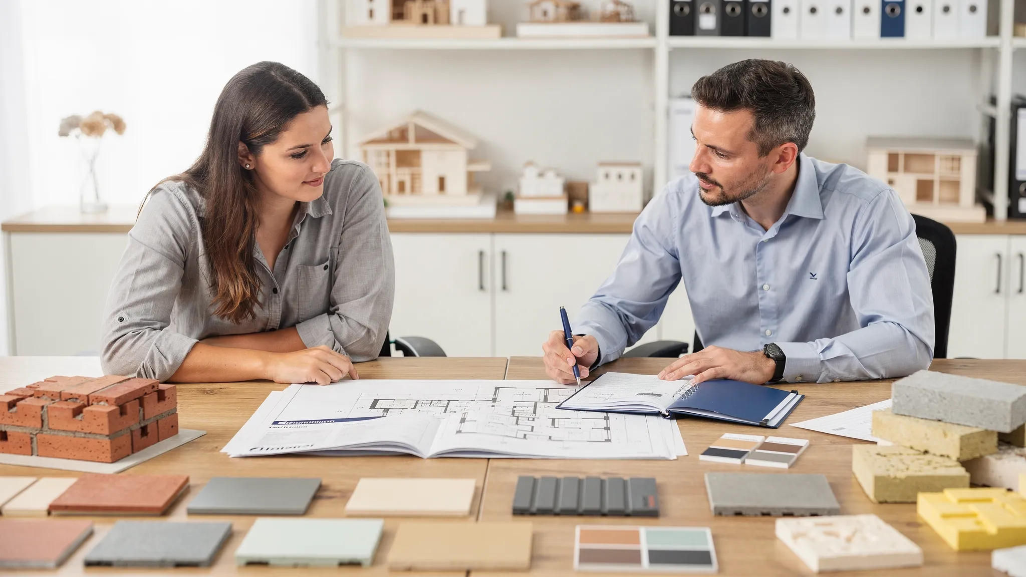 Een koppel dat samen met een architect aan een grote tafel een grondplan en een lastenboek bekijkt, met stalen van gevelmaterialen en isolatie op de voorgrond.