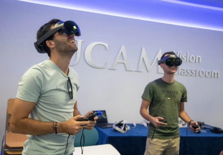 Two individuals are experiencing virtual reality using headsets and controllers inside a modern classroom at European UCAM University, with the institution's logo displayed on the wall in the background.