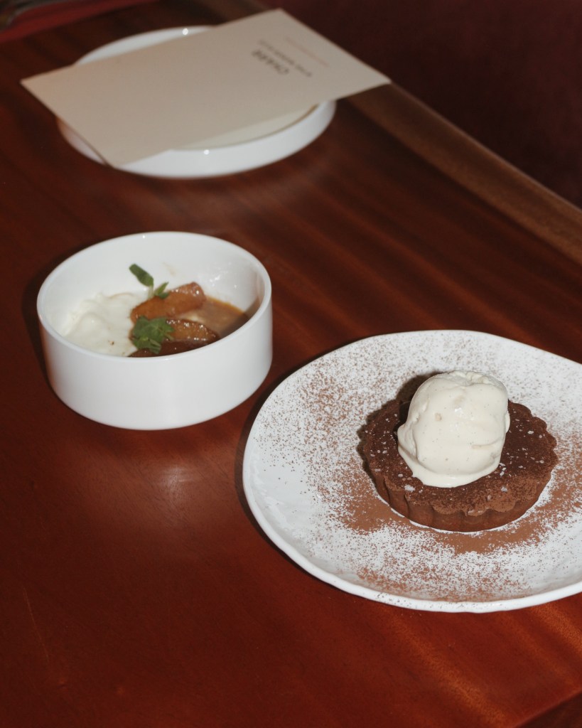 A dessert plate featuring a chocolate tart with a scoop of ice cream on top, alongside a small bowl of caramelized fruit and cream, placed on a wooden table.
