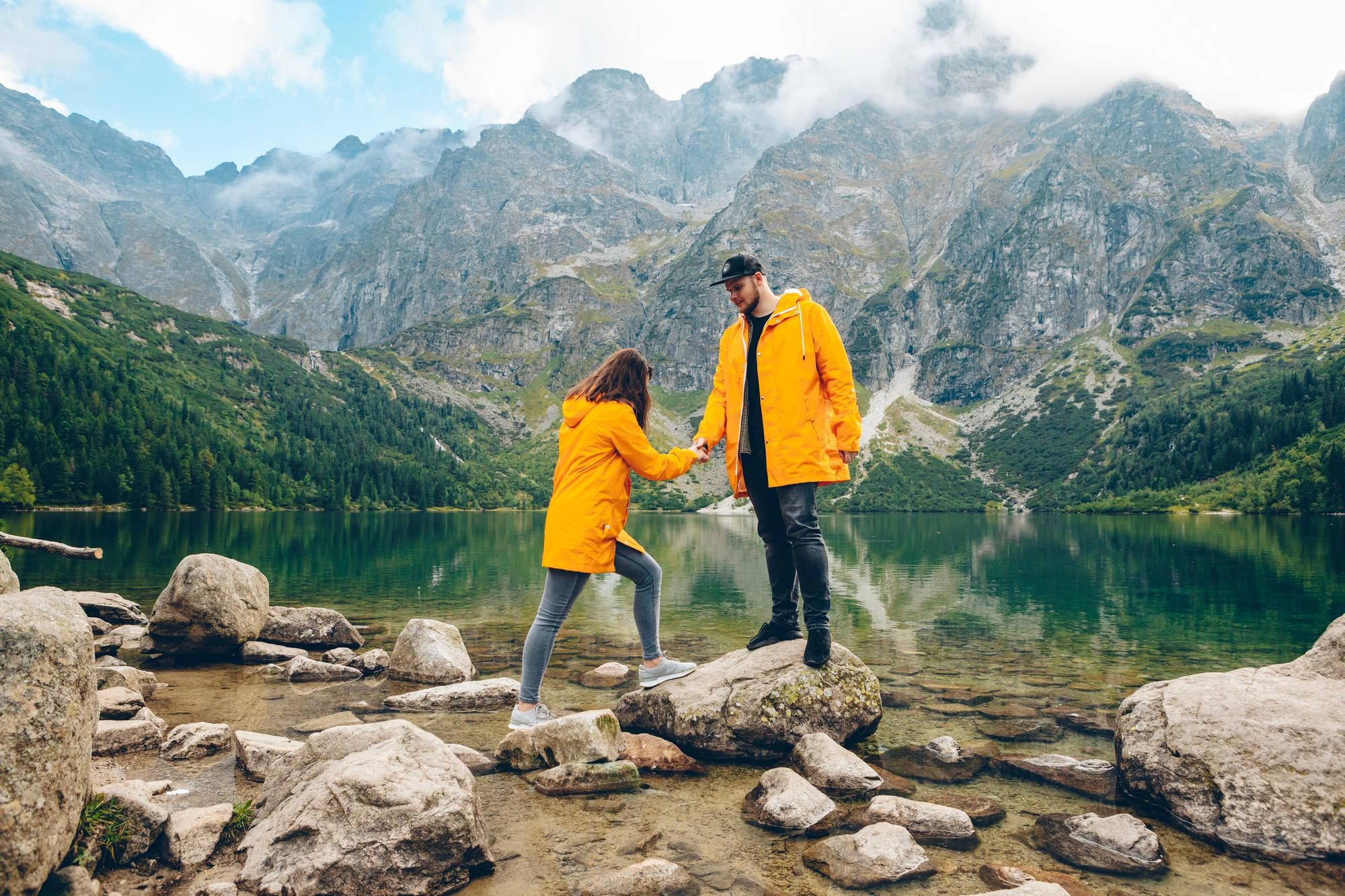 A man in an orange jacket helps a woman in a matching yellow raincoat step across rocks on the shore of a clear mountain lake surrounded by high peaks.
