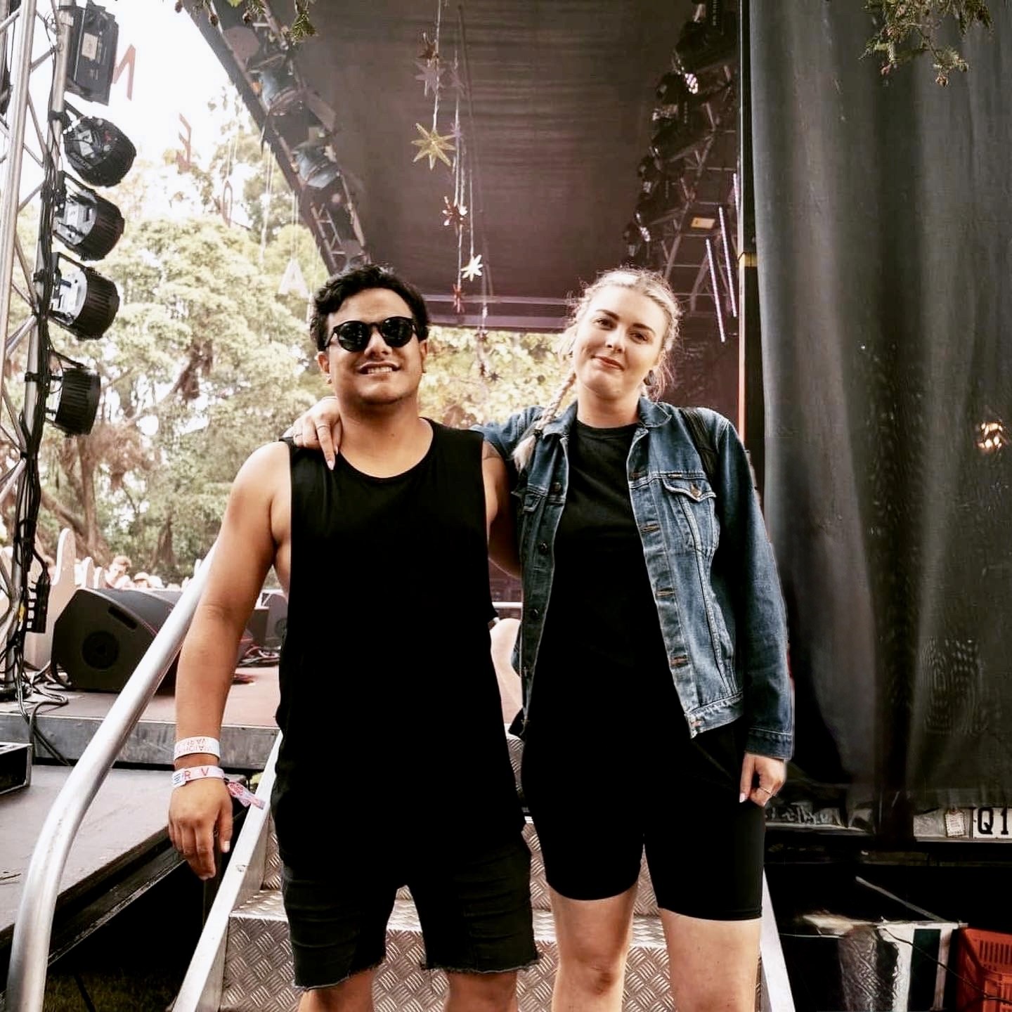 A couple stands on a sandy hill at sunset, facing away, with arms raised in a relaxed "hang loose" gesture. The scene conveys tranquility and joy.