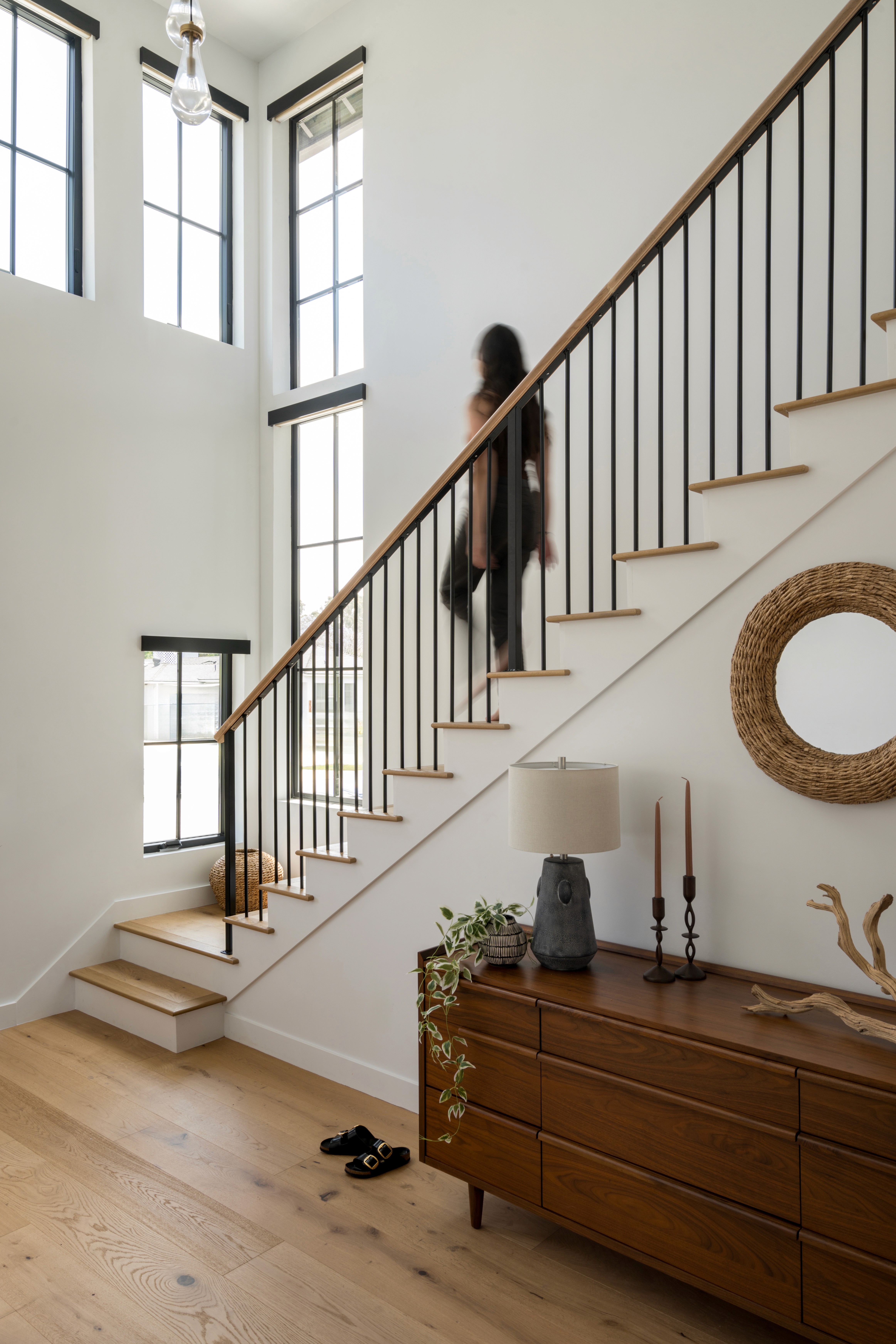 A sleek, modern staircase with minimalist railings and wooden steps leads to the second floor. Overhead, a striking chandelier with cascading lights creates a dramatic focal point, adding elegance and a touch of luxury to the transitional space by Jubilee Interiors in Lakewood, California