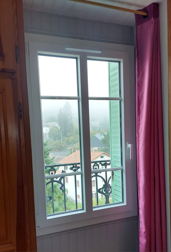 Fenêtre à petits carreaux ouverte sur un balcon avec rideau rose et vue embrumée sur des maisons et de la végétation.