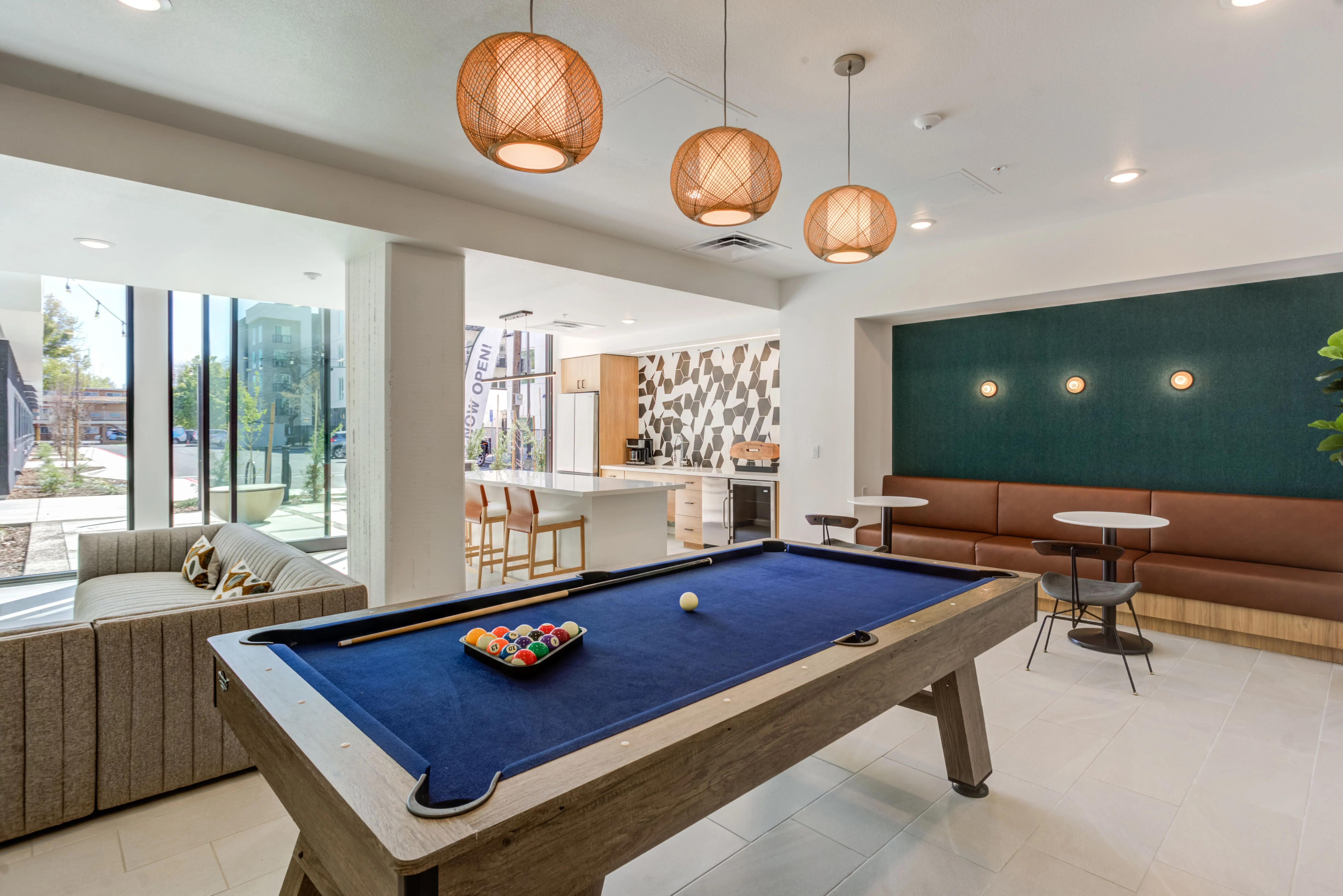 A modern recreational room in Midtown Sacramento featuring a billiards table with a blue felt top, seating area with couch and stools, hanging wicker pendant lights, and a kitchenette with a white countertop. Large windows allow natural light to flood the space, creating a sun-drenched atmosphere.