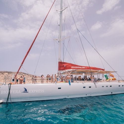 A white sailboat with "BIOSFERA EXPRESS" written on the side, anchored in clear blue water with people on deck under a red canopy.
