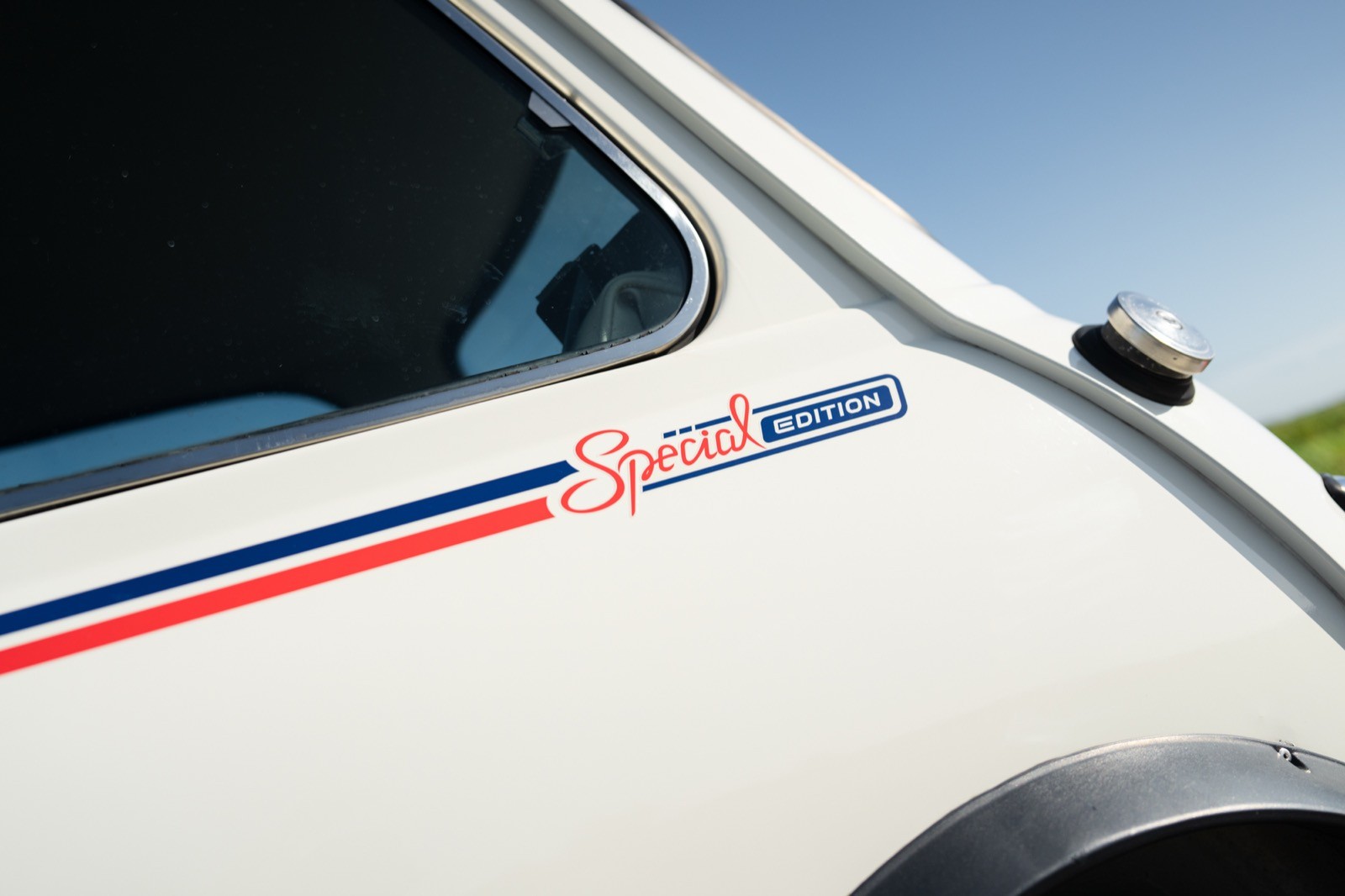 Close-up of a white vehicle’s side, featuring a colorful stripe and a small emblem. Clear blue sky in background.