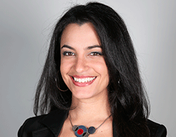 A woman with long, dark hair and dark eyebrows smiles at the camera. She wears a black jacket and a necklace with a circular pendant featuring a red and blue design. Her teeth are visible as she smiles. The background is a plain light gray.