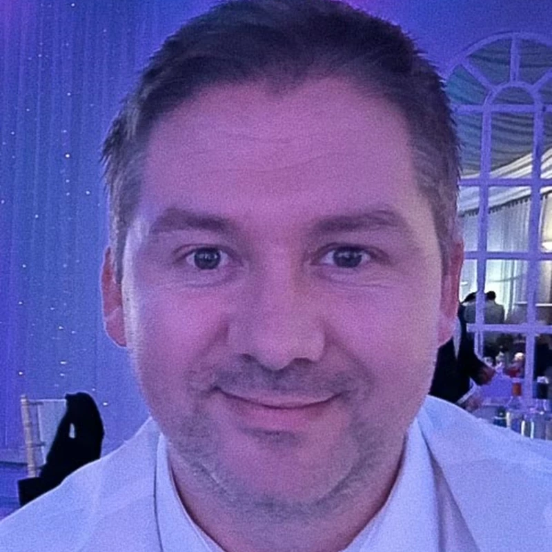 A person wearing a white shirt is seated at an indoor event with blue and purple lighting. Decorative drapes and arched windows create an elegant background.