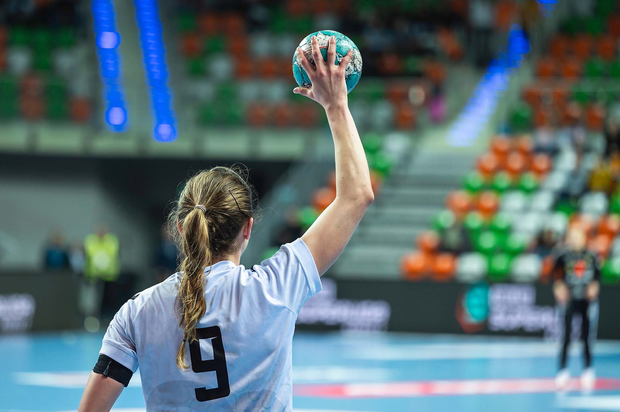 Female handball player in white jersey number 9 raises ball overhead preparing to throw during an indoor match.