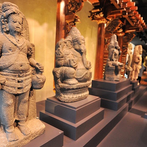 A row of ancient stone statues on pedestals in a museum, with intricate carved wooden columns in the background.