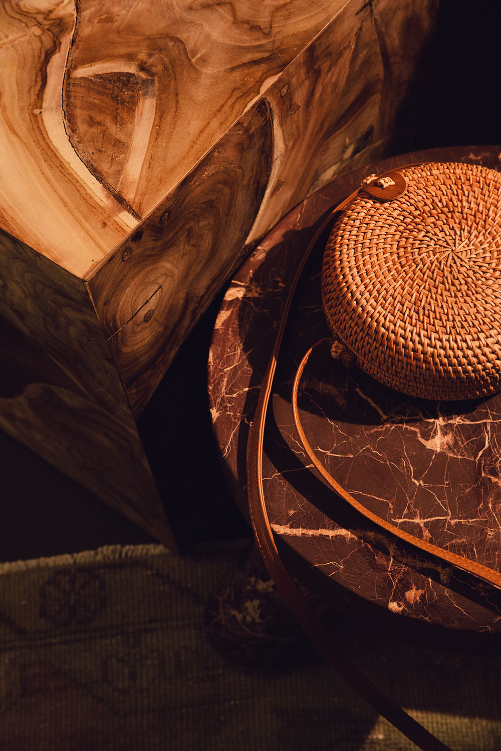 A warm tonned, wood grained coffee table next to a marble bench. A wicker handback rests on the bench with looping leather straps