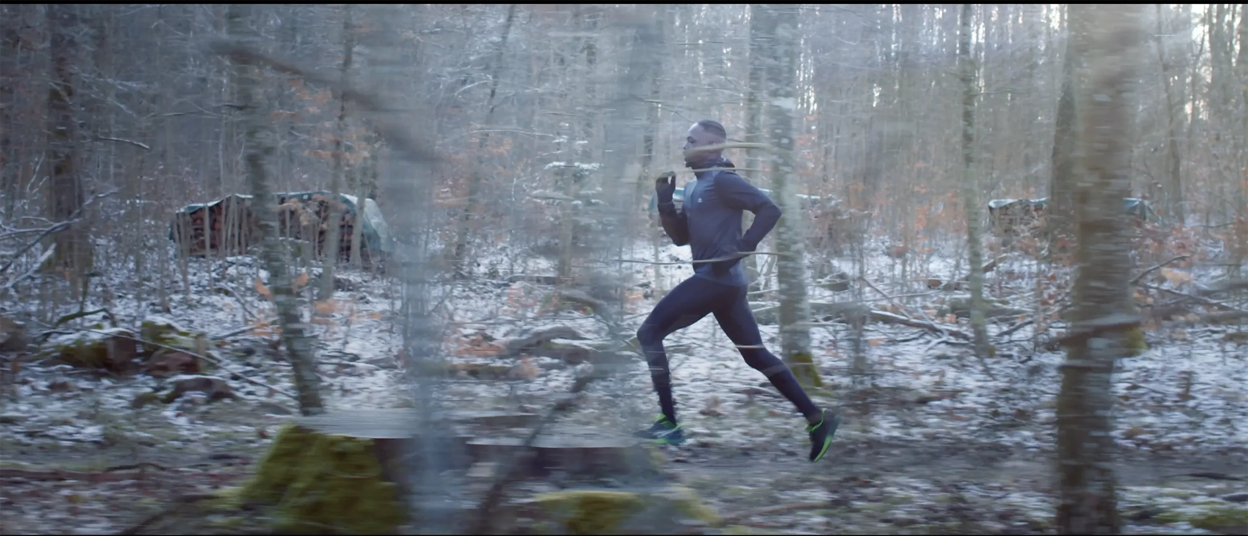 A wide cinematic shot of a man running through a snow dusted winter forest with soft natural lighting.