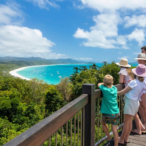 四個人站在一個木製瞭望台上,凝望著一個擁有碧綠海水、船隻和茂盛綠色背景的熱帶沙灘。