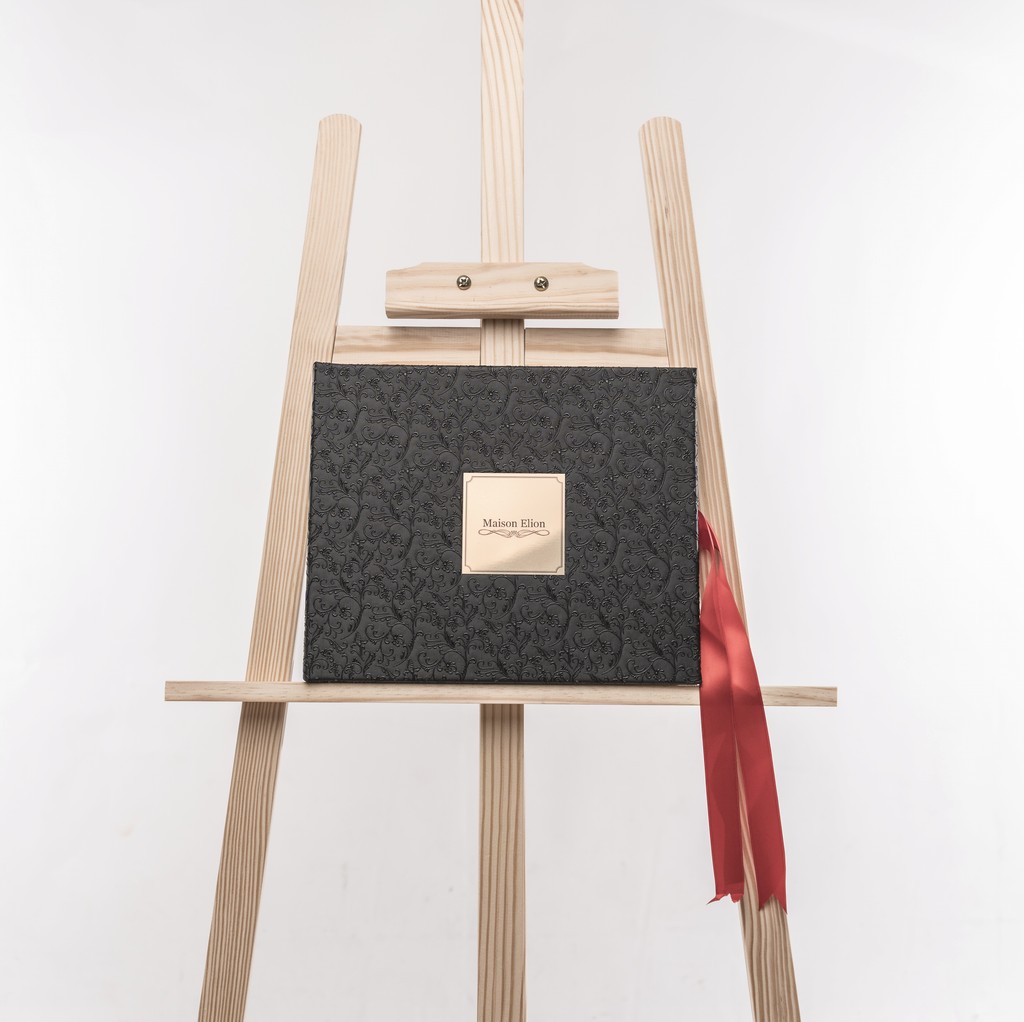 Black embossed Fine Art album adorned with a gold Maison Elion plaque and red ribbon — the union of gravity and desire. Album Fine Art noir embossé, orné d’une plaque dorée Maison Elion et d’un ruban rouge — l’union de la gravité et du désir.