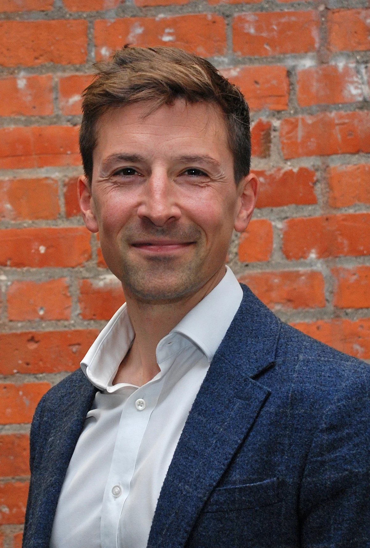 A person wearing a white shirt and blue blazer stands in front of a red brick wall.