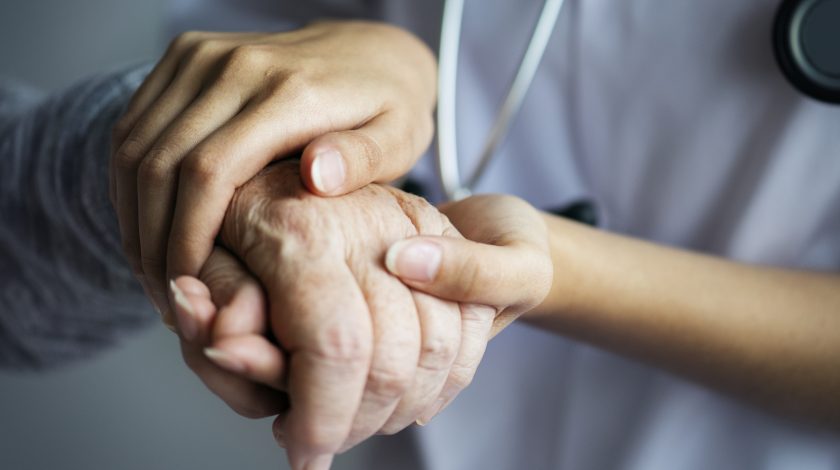 A close-up image shows a caregiver gently holding the hand of an elderly person, symbolizing compassion and support in healthcare settings.