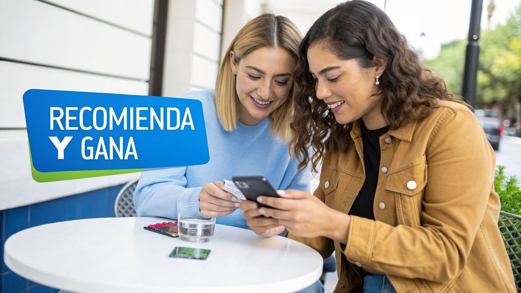Dos mujeres sonrientes mirando un celular en una mesa con un cartel 'RECOMIENDA Y GANA'.