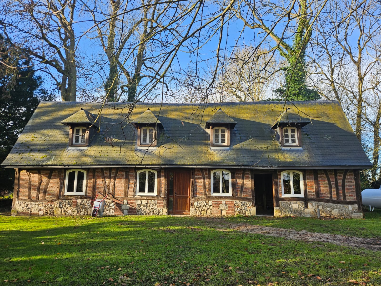 Traditional Normandy brick-and-timber farmhouse surrounded by trees, serving as the Parcel Torrefaction coffee roastery location, with moss-tinted slate roof and garden in front.