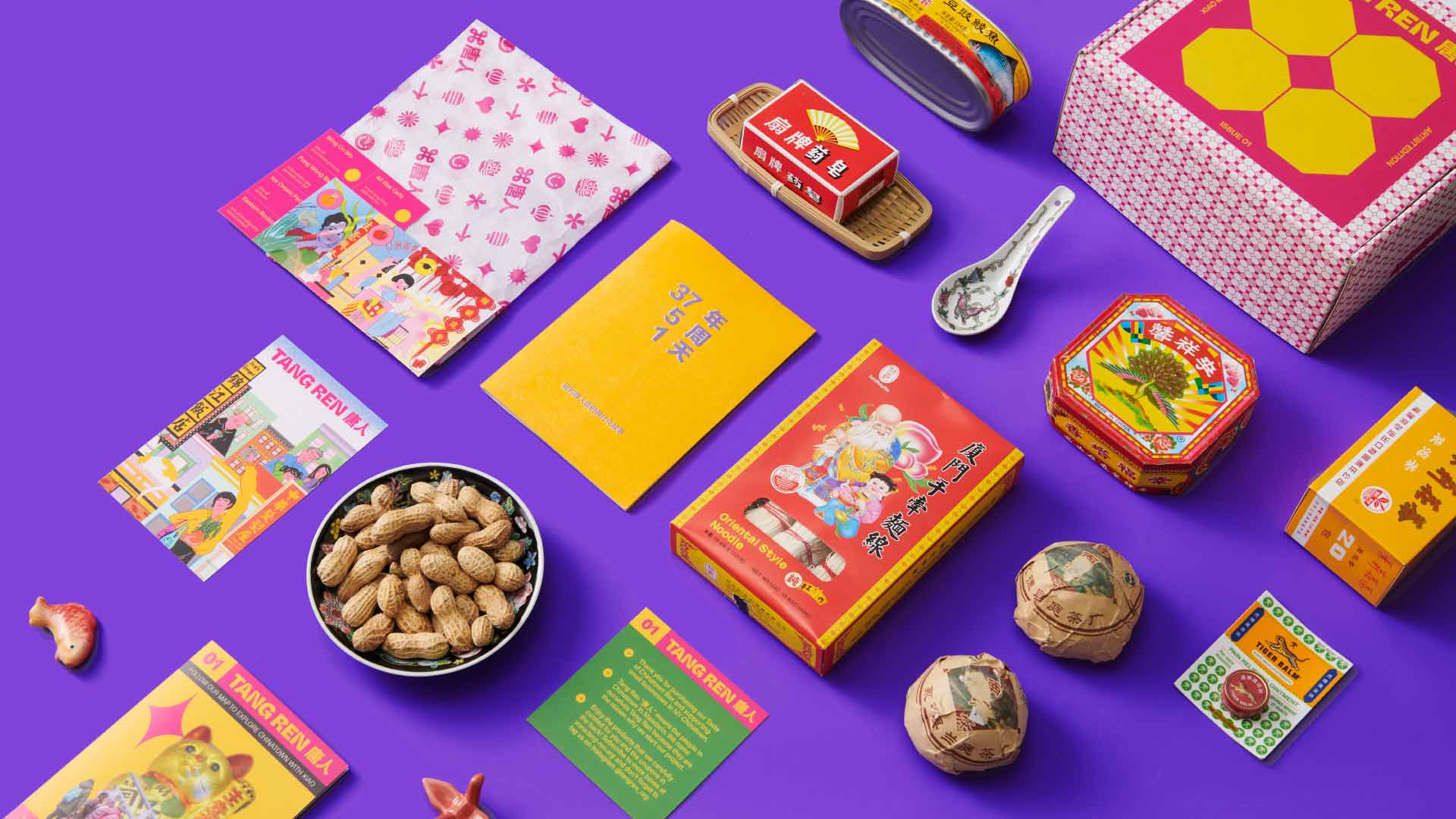 The image shows a collection of items arranged on a vibrant purple background. These include two books, one yellow and one orange, with Chinese characters printed on them, a small white spoon, a red tin with a yellow lid, a box with a yellow lid and a red and white pattern, a box with a yellow lid and a red and white pattern, a small white rectangular packet with green text, a small orange rectangular packet with green text, a black bowl filled with peanuts, a small red figurine, a small orange figurine, a small green rectangular packet with text, two small brown objects that appear to be made of paper or cardboard, and a small red and white box with a yellow design.