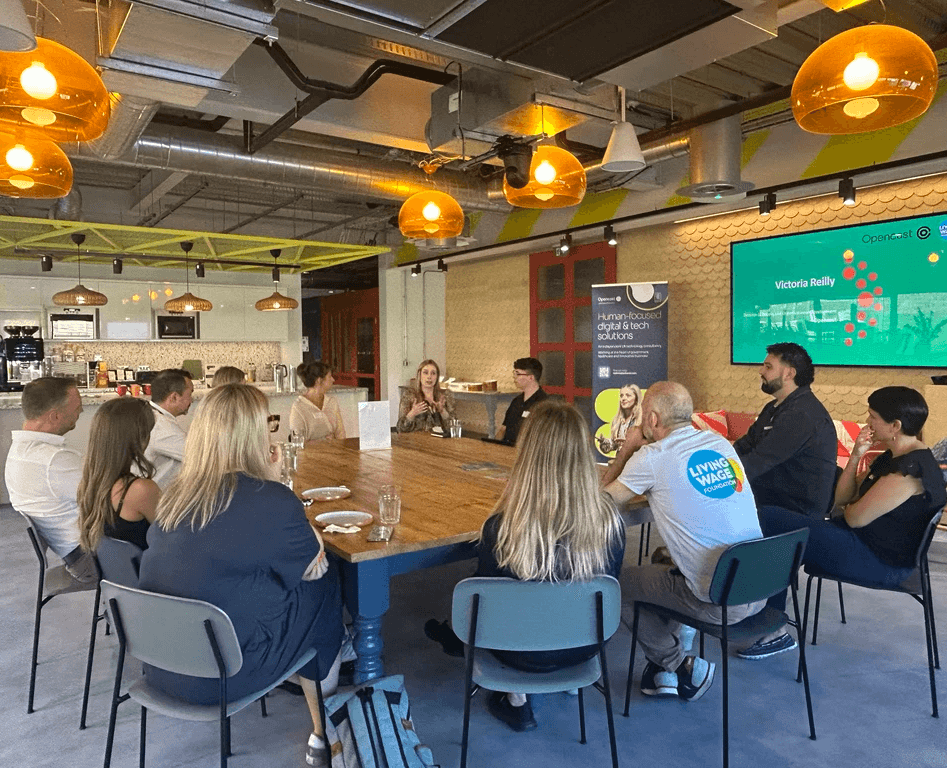 A group of around a dozen people sit around a large wooden table in a modern meeting space, listening to a speaker at the far end. The room features a café-style kitchen area, exposed ceilings, and large amber pendant lights. A presentation slide is displayed on a screen beside an Opencast banner, creating an informal networking or discussion event setting.