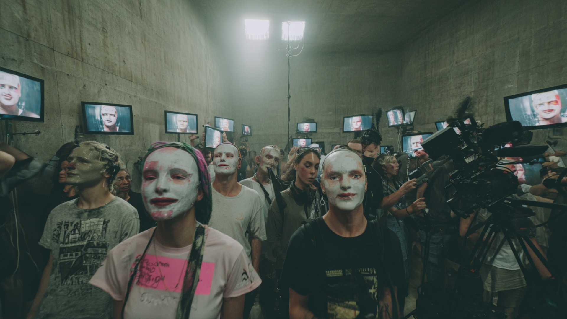 A cinematic, wide-angle photo in the style of Stanley Kubrick showing rows of uncanny, zombie-like influencers in trendy streetwear standing in a stark concrete warehouse. They stare blankly at a massive wall of screens playing bright, synthetic AI video content, representing the 'Dead Internet' theory.