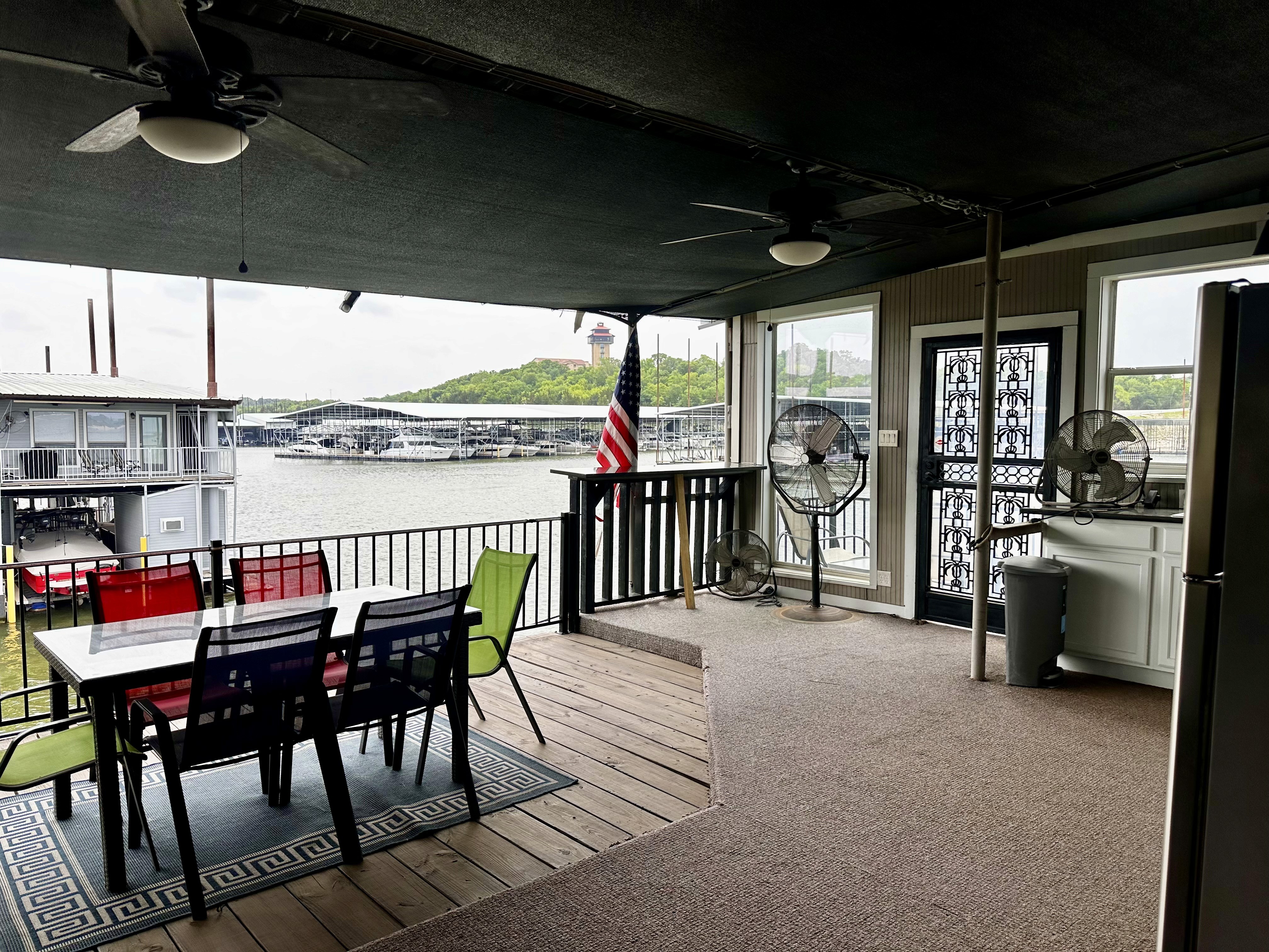 Covered outdoor seating area overlooking a lake with a table, chairs, fans, and an American flag.