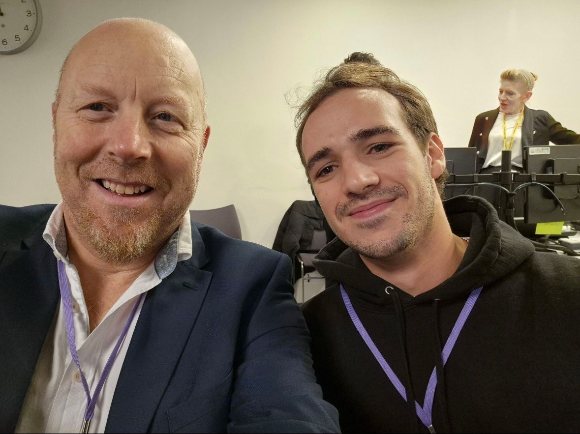 Two men at the Digital Planning Directory Launch smiling for a selfie, wearing lanyards in a conference setting