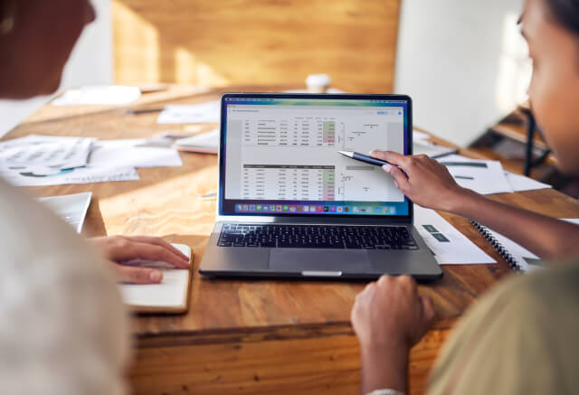 Two people sit at a wooden table, reviewing financial data and charts displayed on a laptop screen, with documents and a notebook nearby.