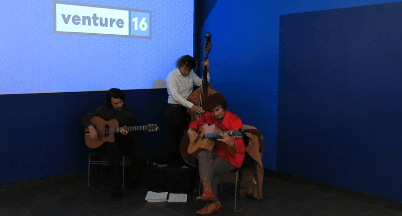A group of students performing on stage at various musical instruments, while standing in front of a blue projector screen with text logo: venture 16.