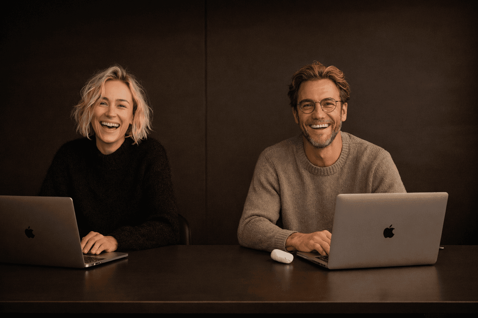Two people sit at a dark wooden table, smiling warmly at the camera. They have laptops open, creating a friendly and collaborative atmosphere.