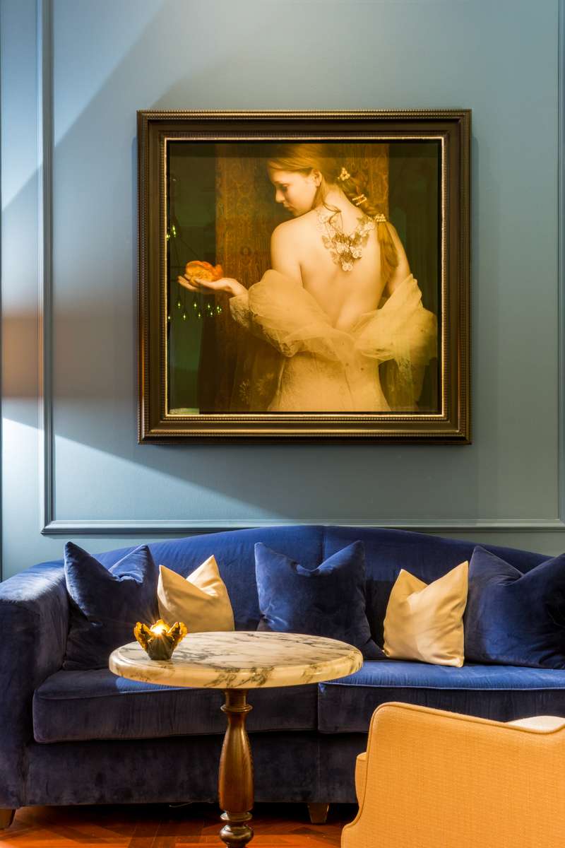 A framed painting of a woman in a green dress hangs above a blue velvet sofa with gold and cream pillows.
