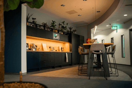 Moderne Büroküche mit dunklen Schränken, beleuchtetem Regal, Pflanzen auf dem Oberschrank und hohem Tisch mit Stühlen