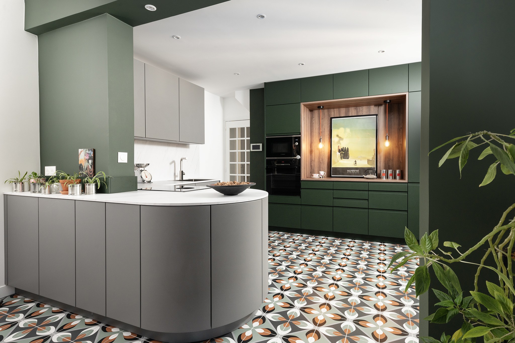 Cuisine moderne avec armoires vertes et grises, carrelage à motifs, four encastré, œuvre d'art encadrée, plantes en pot sur le comptoir et éclairage chaleureux, avec une plante feuillue au premier plan.