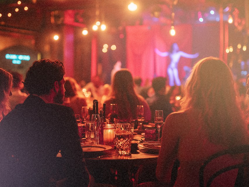 Vue d'un dîner dans un cabaret avec des invités assis autour de tables élégantes, regardant une performance sur scène, éclairée par des lumières tamisées.