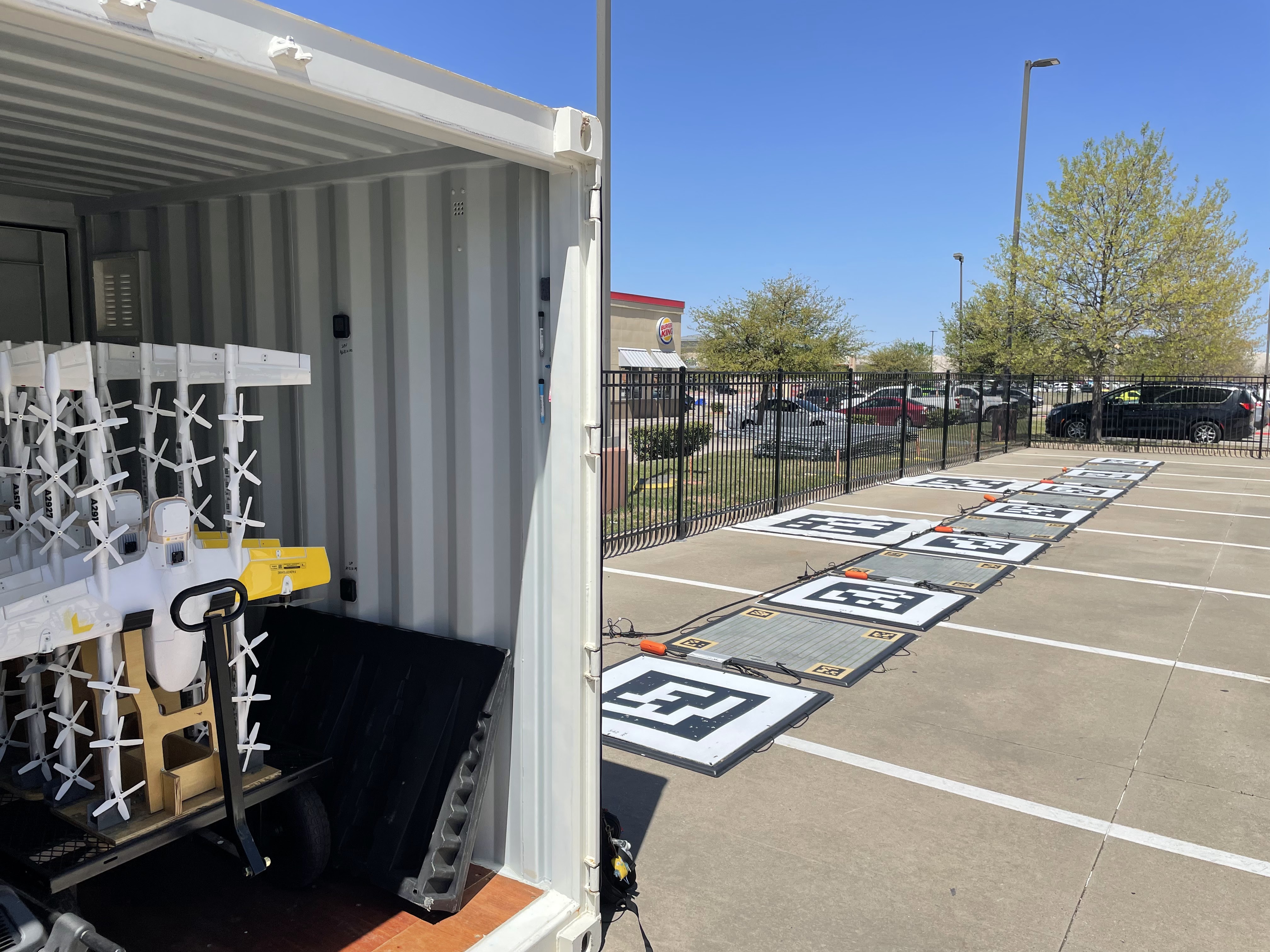 A shipping container with industrial equipment is open on a sunny day next to a parking lot, featuring marked square platforms lined up along a fenced area, with several trees visible in the background under a clear blue sky.