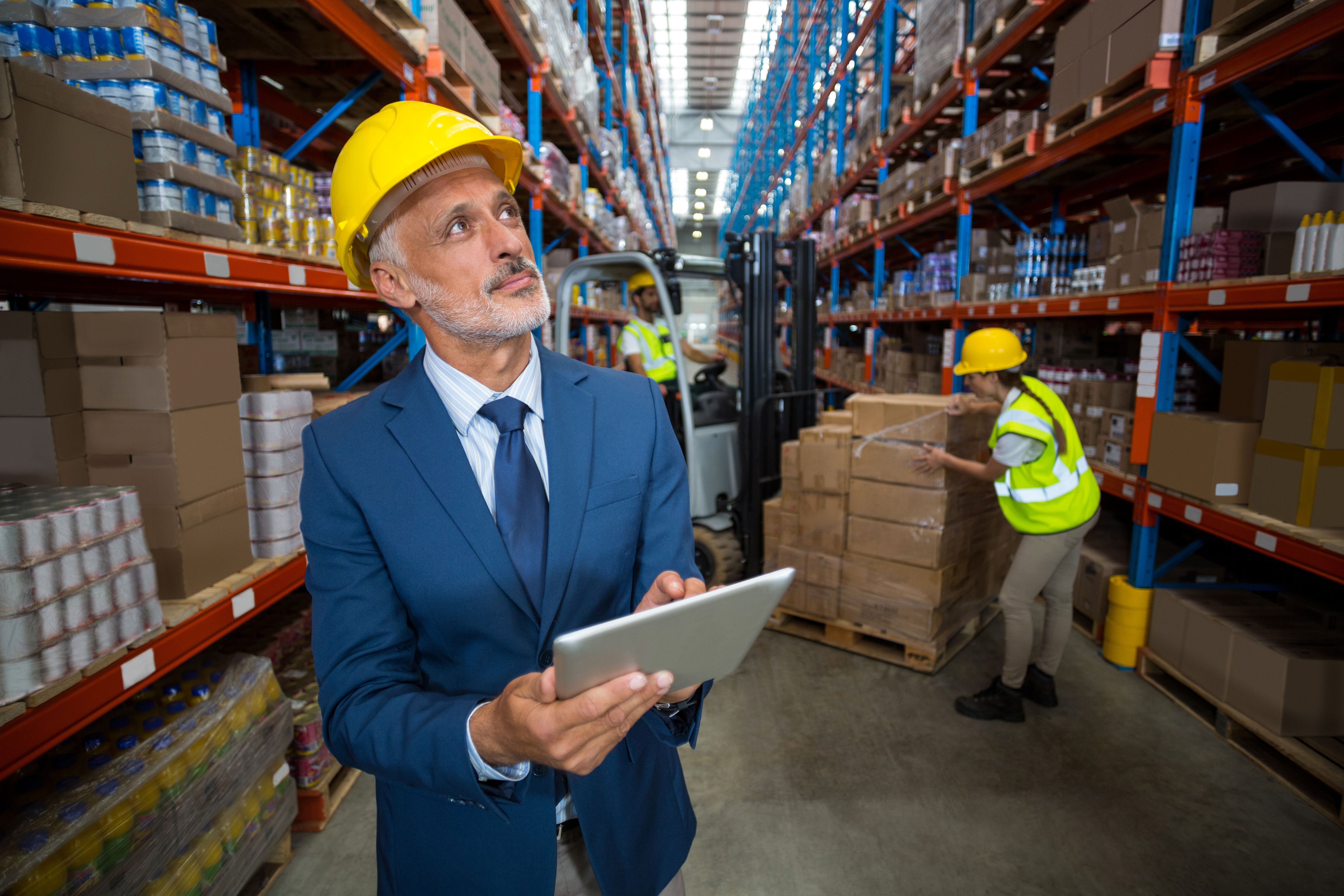 A man in blue suit wearing yellow safety helmet using a tablet device working in a warehouse