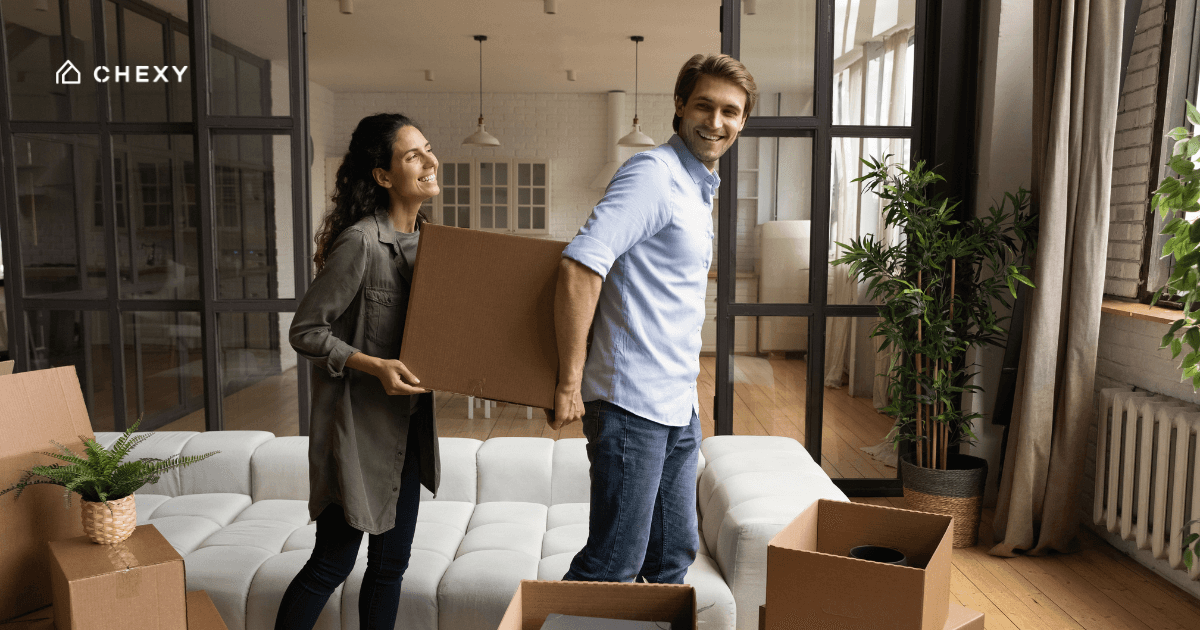 A couple carrying a moving box in a half-furnished apartment