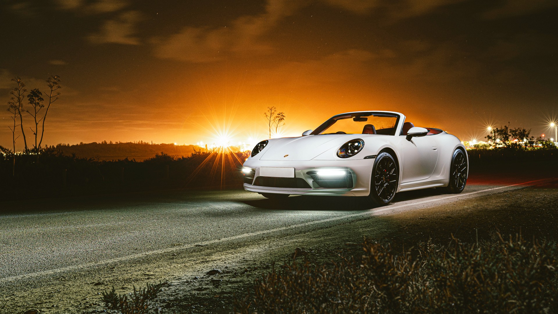 White Porsche 911 (992) Cabriolet with black wheels and roof down, photographed from the front three-quarter at night, headlights and front light bar lit, with a sunburst flare and warm orange afterglow on the horizon behind.