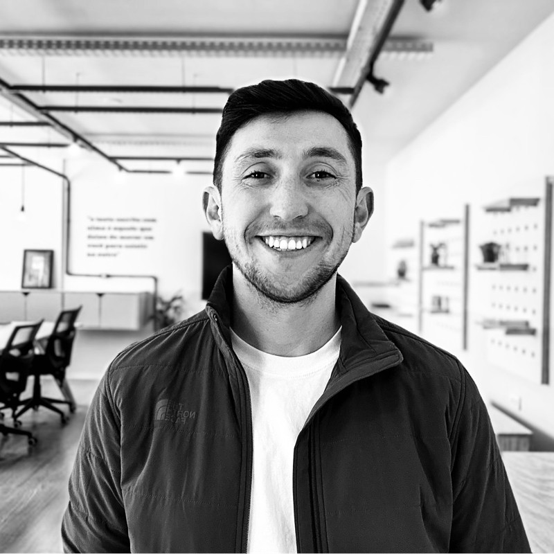 Landon in a black and white photo stands in a modern indoor space with blurred furniture in the background.