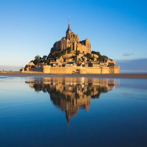 A historic island abbey and village reflected in calm water under a clear blue sky.