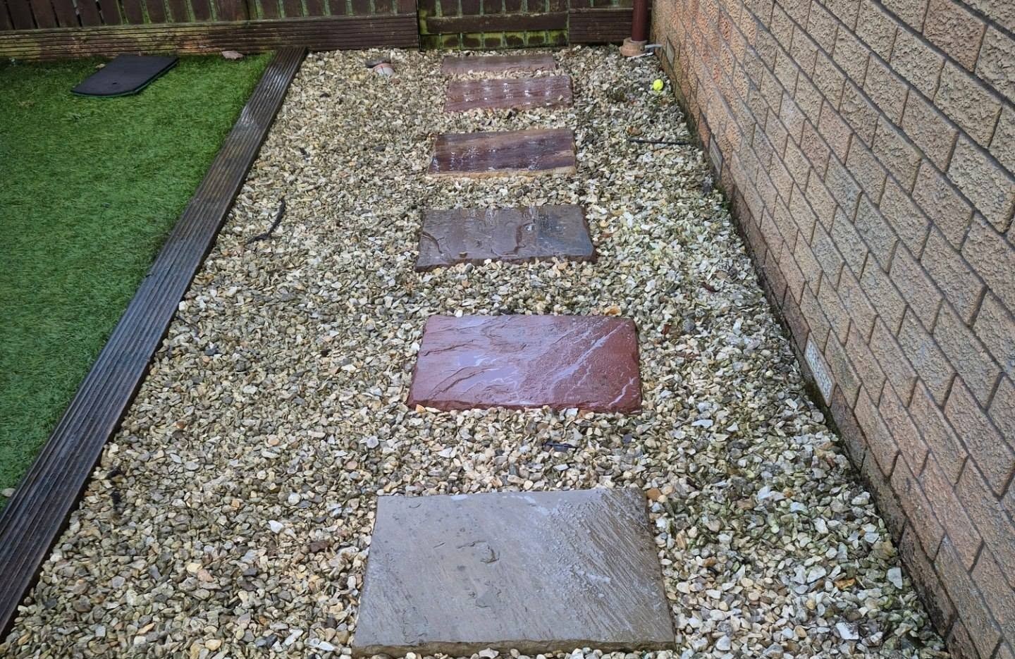 A garden path with five stepping stones set into gravel, bordered by grass on the left and a brick wall on the right.