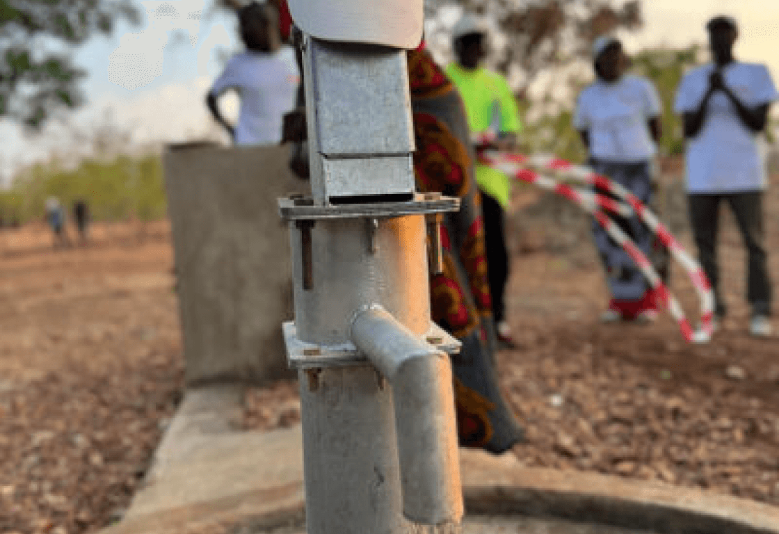 Close-up of a clean water well pump installed by Osmow's Hope Fund in a developing community with local residents in the background